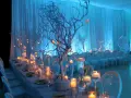 A long table set for an event with white chairs, candles, and white tree branches decorated with small lights.