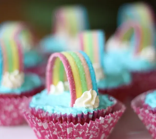 Sju cupcakes med ljusblå regnbågs frosting och godisregnbågar. Rosa muffinsformar med mönster.