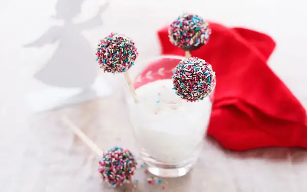 Söta cake pops med pepparkakssmak, dekorerade med färgglada strössel. En vit ängel i bakgrunden.