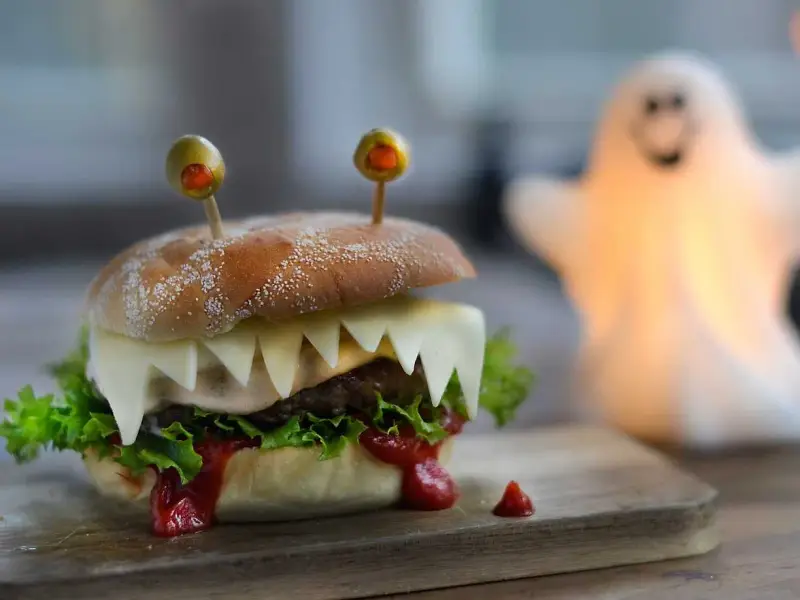 En läskig halloween hamburgare med osttänder och olivögon. En spöklampa lyser upp i bakgrunden.