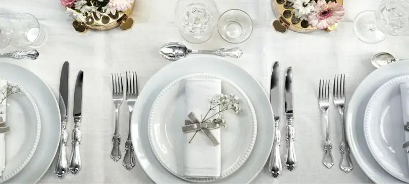 Ett elegant dukat bord med vita tallrikar, servetter dekorerade med blommor och vackra, silverfärgade engångsbestick.